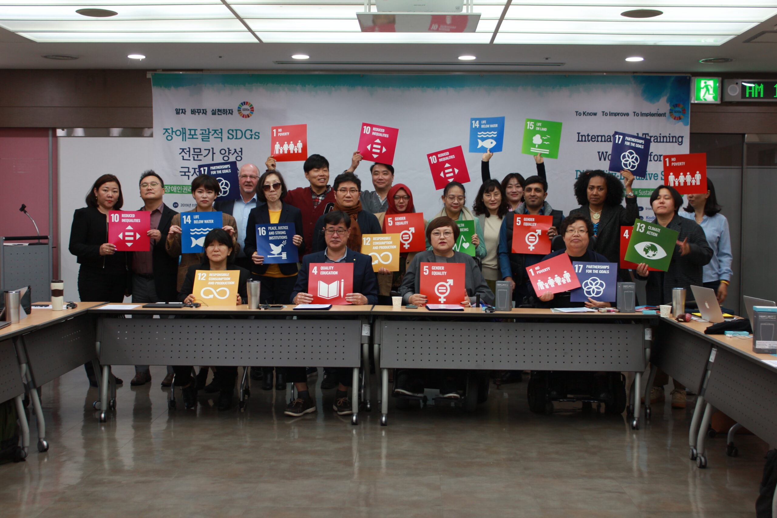 Delegates at the Korean Disability Forum Workshop in November 2019, seated and standing behind desks holding up cards which have prined on them the Sustainable Development Goals.